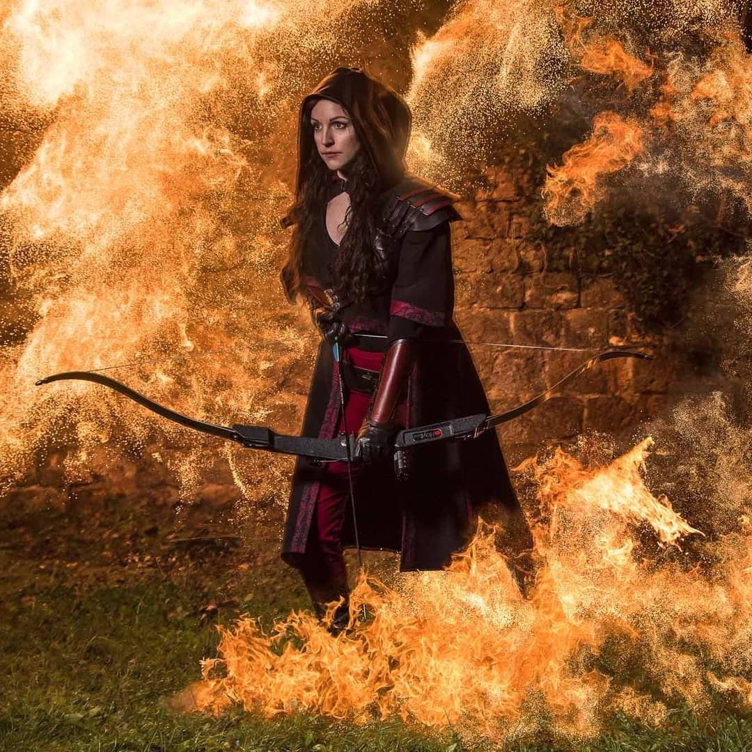 Archère en costume, flèche pointée, mur de flammes dans le dos